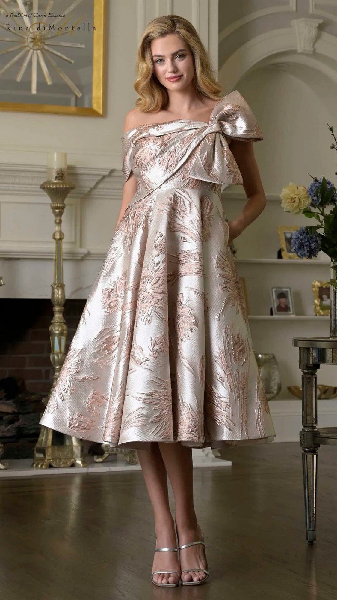 A woman stands indoors wearing the Rina Di Montella RD207 knee-length, mauve multi semi-formal dress with an off-the-shoulder design, jacquard floral patterns, and a large bow detail, paired with strappy heels.